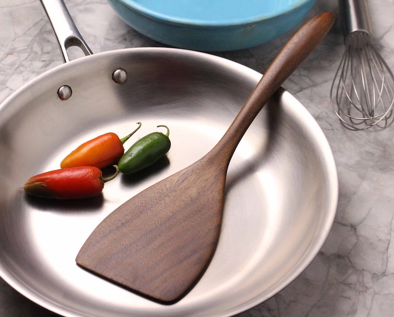 May include: A stainless steel frying pan with a wooden spatula and three chili peppers. The spatula is dark brown and has a wide, flat head. The peppers are red, orange, and green.