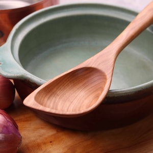 May include: A wooden spoon rests in a ceramic bowl with a green interior and brown exterior. Two red onions are on a wooden surface. A copper pot is in the background. The spoon is light brown and has a curved handle.