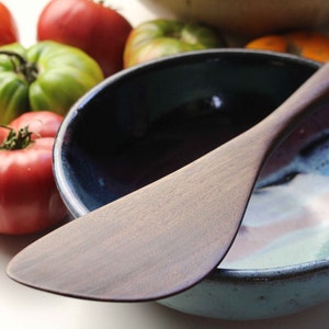 May include: A wooden spatula lies across a ceramic bowl with a blue and teal interior. Ripe red and green tomatoes are scattered around the bowl, suggesting a kitchen setting. The spatula has a smooth, light brown finish.