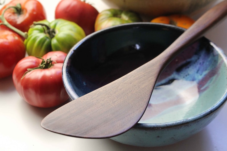 May include: A wooden spatula lies across a ceramic bowl with a blue and teal interior. Ripe red and green tomatoes are scattered around the bowl, suggesting a kitchen setting. The spatula has a smooth, light brown finish.