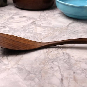 May include: A wooden spoon with a curved handle and a wide, flat head. The spoon is made of a dark brown wood, and the grain is visible. The spoon is approximately 12 inches long, and is designed for cooking and serving.