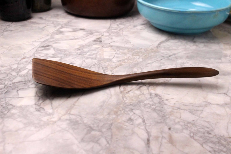 May include: A wooden spoon with a curved handle and a wide, flat head. The spoon is made of a dark brown wood, and the grain is visible. The spoon is approximately 12 inches long, and is designed for cooking and serving.