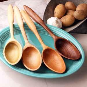 May include: Four wooden spoons in shades of light and dark brown are arranged on a turquoise plate. The spoons are displayed in a row, highlighting their different wood grains. Potatoes and garlic are in the background.