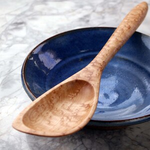May include: A wooden spoon with a light-colored, patterned handle rests on a blue ceramic bowl. The bowl has a dark rim and a glossy finish. The spoon's bowl shape is oval. The background is a marble-like surface.