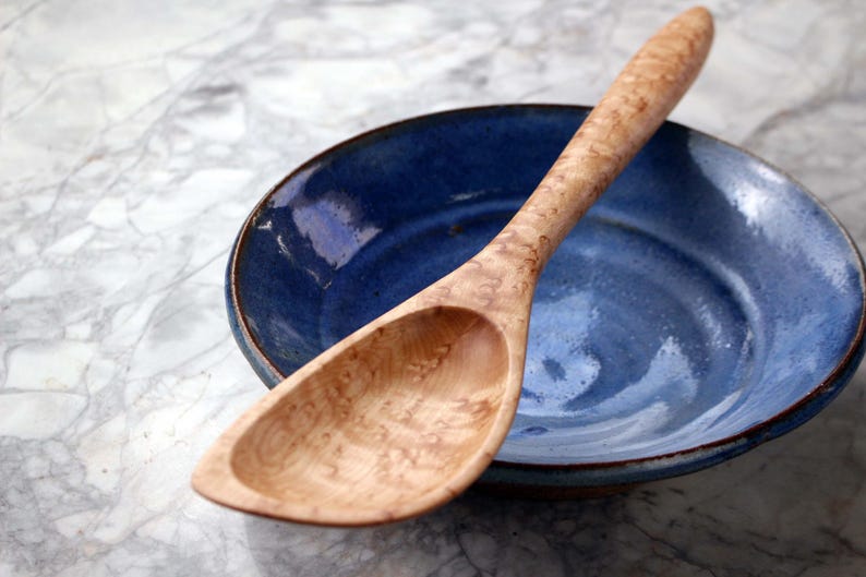 May include: A wooden spoon with a light-colored, patterned handle rests on a blue ceramic bowl. The bowl has a dark rim and a glossy finish. The spoon's bowl shape is oval. The background is a marble-like surface.