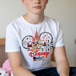 Op de afbeelding: Wit T-shirt met een kleurrijke Disney-afbeelding. Het ontwerp bevat het Disney-logo, een kasteel en verschillende stripfiguren. De afbeelding wordt omlijst door Mickey Mouse oren en vuurwerk. De tekst "Disney 2026" is ook zichtbaar.