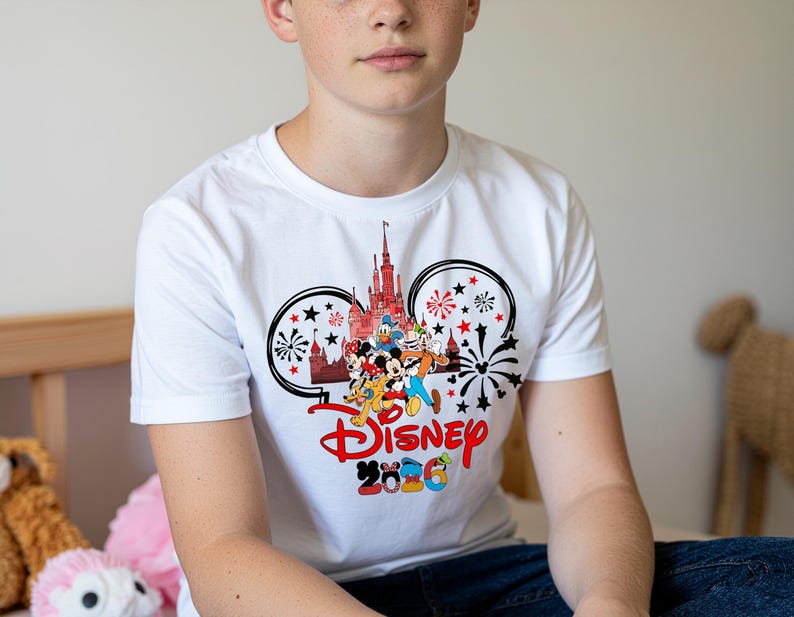 Op de afbeelding: Wit T-shirt met een kleurrijke Disney-afbeelding. Het ontwerp bevat het Disney-logo, een kasteel en verschillende stripfiguren. De afbeelding wordt omlijst door Mickey Mouse oren en vuurwerk. De tekst "Disney 2026" is ook zichtbaar.