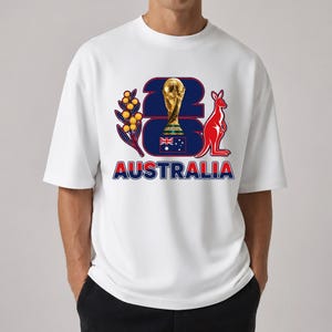 Puede incluir: Camiseta blanca con un dise&ntilde;o gr&aacute;fico que presenta el n&uacute;mero '23' en azul marino, un trofeo dorado, un canguro rojo y la palabra 'AUSTRALIA' en rojo y azul. El dise&ntilde;o incluye una ramita con bayas amarillas.