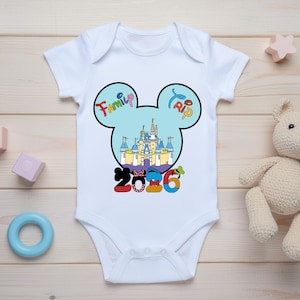 Op de afbeelding: Witte babybody met een kleurrijk Mickey Mouse-ontwerp. Het ontwerp toont een kasteel, de woorden "Family Trip" en het jaar "2016". Speelgoed omringt de body.