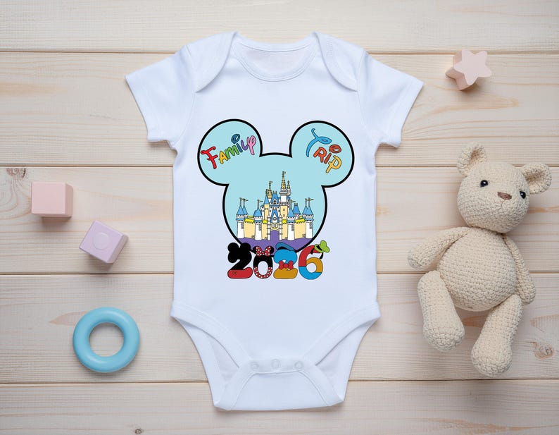 Op de afbeelding: Witte babybody met een kleurrijk Mickey Mouse-ontwerp. Het ontwerp toont een kasteel, de woorden "Family Trip" en het jaar "2016". Speelgoed omringt de body.
