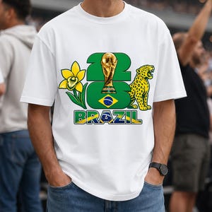 Puede incluir: Camiseta blanca con un dise&ntilde;o que incluye el n&uacute;mero "23", un trofeo dorado, una flor amarilla y un jaguar. La palabra "BRAZIL" est&aacute; impresa en verde y amarillo. El dise&ntilde;o incorpora los colores de la bandera brasile&ntilde;a.