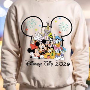 May include: A cream-colored sweatshirt featuring a Disney Trip 2026 graphic. The graphic includes Mickey Mouse, Minnie Mouse, Donald Duck, Goofy, Pluto, and a castle. The design is framed by a Mickey Mouse ear outline with fireworks.
