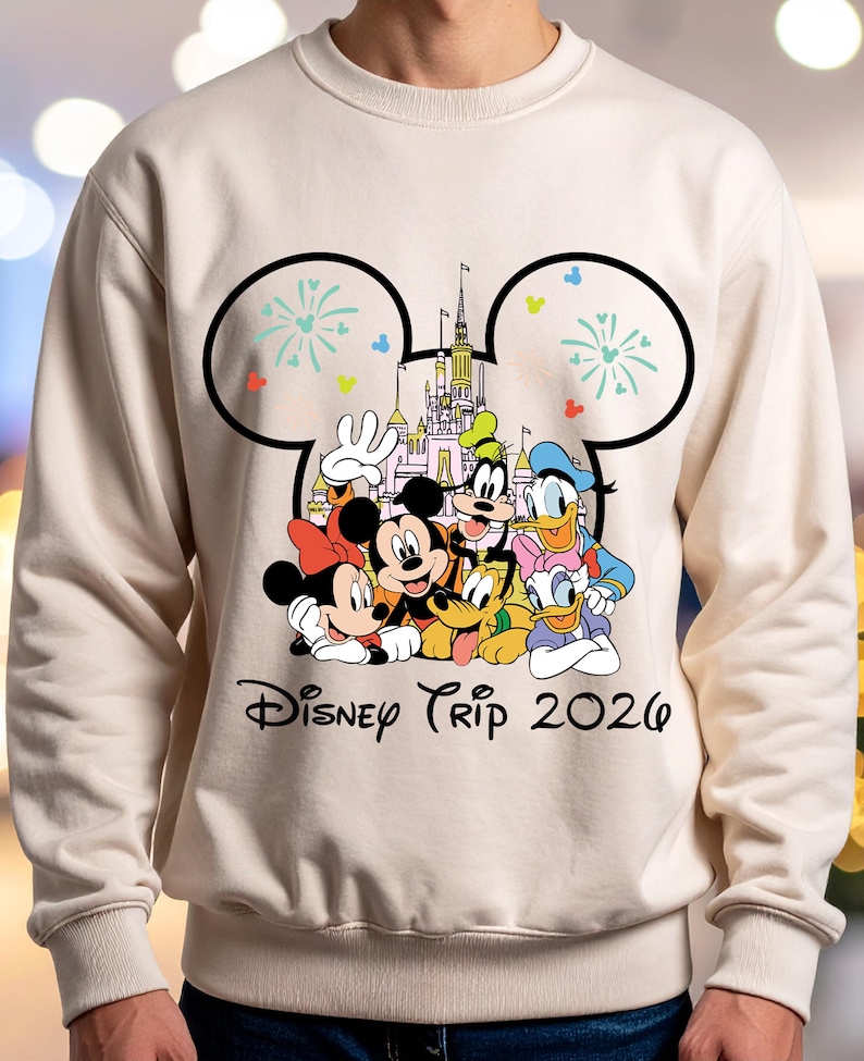 May include: A cream-colored sweatshirt featuring a Disney Trip 2026 graphic. The graphic includes Mickey Mouse, Minnie Mouse, Donald Duck, Goofy, Pluto, and a castle. The design is framed by a Mickey Mouse ear outline with fireworks.