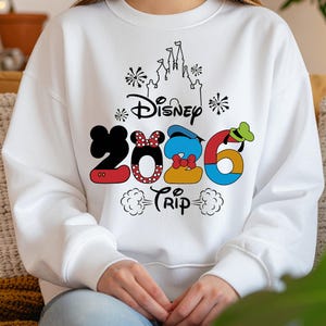 Op de afbeelding: Witte sweatshirt met een kleurrijke Disney 2026 Trip-afbeelding. Het ontwerp bevat Mickey Mouse-oren, Minnie Mouse's strik, Donald Duck's hoed en Goofy's hoed in de cijfers. Een kasteelsilhouet en vuurwerk maken het ontwerp compleet.