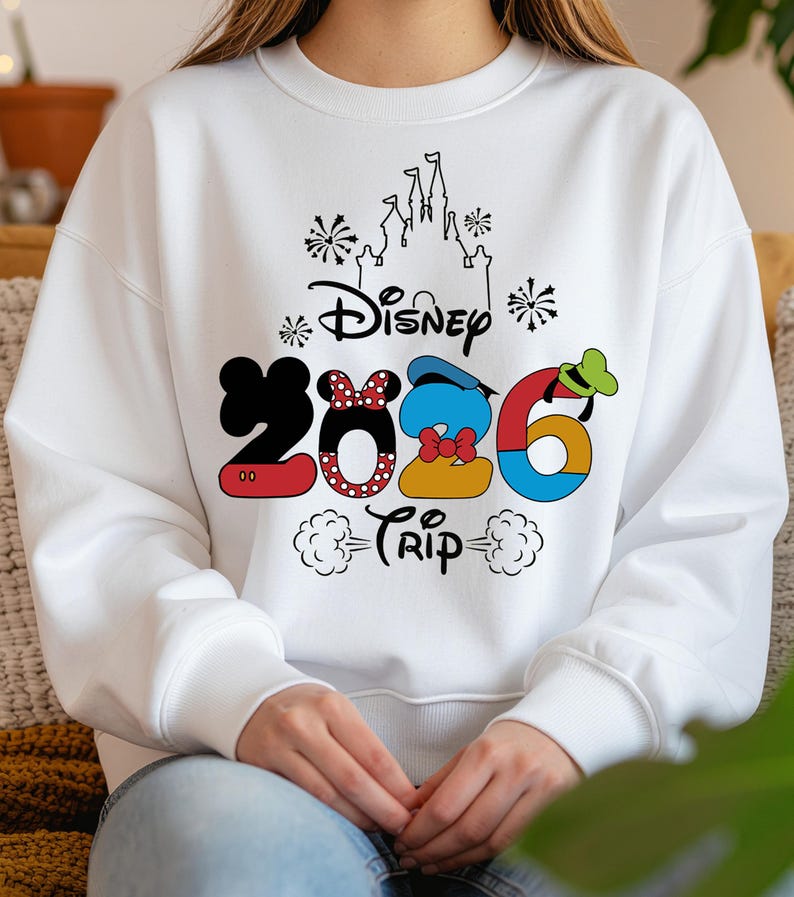 Op de afbeelding: Witte sweatshirt met een kleurrijke Disney 2026 Trip-afbeelding. Het ontwerp bevat Mickey Mouse-oren, Minnie Mouse's strik, Donald Duck's hoed en Goofy's hoed in de cijfers. Een kasteelsilhouet en vuurwerk maken het ontwerp compleet.