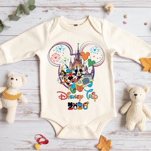 Op de afbeelding: Cr&egrave;me baby-romper met een kleurrijke Disney Trip 2026-afbeelding. Het ontwerp toont Mickey Mouse, Minnie Mouse en andere personages voor een kasteel, met vuurwerk en ballonnen. De romper heeft lange mouwen.