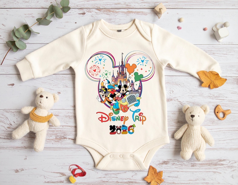 Op de afbeelding: Cr&egrave;me baby-romper met een kleurrijke Disney Trip 2026-afbeelding. Het ontwerp toont Mickey Mouse, Minnie Mouse en andere personages voor een kasteel, met vuurwerk en ballonnen. De romper heeft lange mouwen.