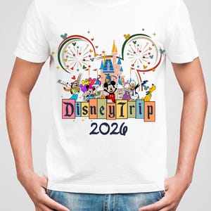 May include: White t-shirt featuring a colorful graphic of Disney characters, a castle, and fireworks. The text "Disney Trip 2026" is displayed below the characters. The design includes Mickey Mouse, Donald Duck, and Goofy.