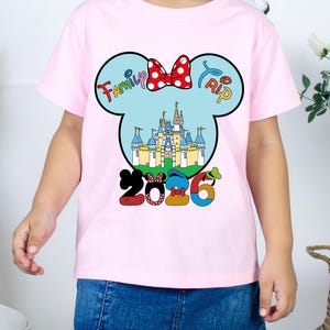 Op de afbeelding: Een roze t-shirt met een Mickey Mouse-hoofdontwerp, een kasteel, een rode strik en de woorden "Family Trip 2026". Het t-shirt wordt door een kind gedragen.