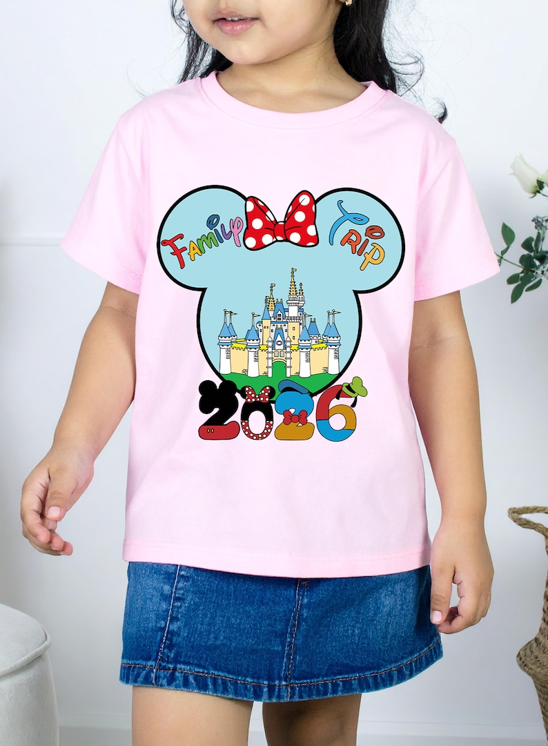 Op de afbeelding: Een roze t-shirt met een Mickey Mouse-hoofdontwerp, een kasteel, een rode strik en de woorden "Family Trip 2026". Het t-shirt wordt door een kind gedragen.