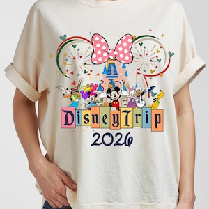May include: Cream-colored t-shirt featuring a Disney Trip 2026 design. The design includes a pink polka-dot bow, a castle, and various Disney characters. The text "Disney Trip" is displayed above the year 2026.