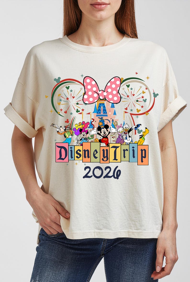 May include: Cream-colored t-shirt featuring a Disney Trip 2026 design. The design includes a pink polka-dot bow, a castle, and various Disney characters. The text "Disney Trip" is displayed above the year 2026.