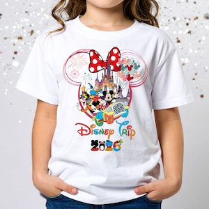 May include: White t-shirt featuring a colorful Disney Trip design. The graphic includes Mickey and Minnie Mouse, Donald Duck, and Goofy in a car, with a castle and fireworks. The design also includes a red bow and the text "Disney Trip 2026".