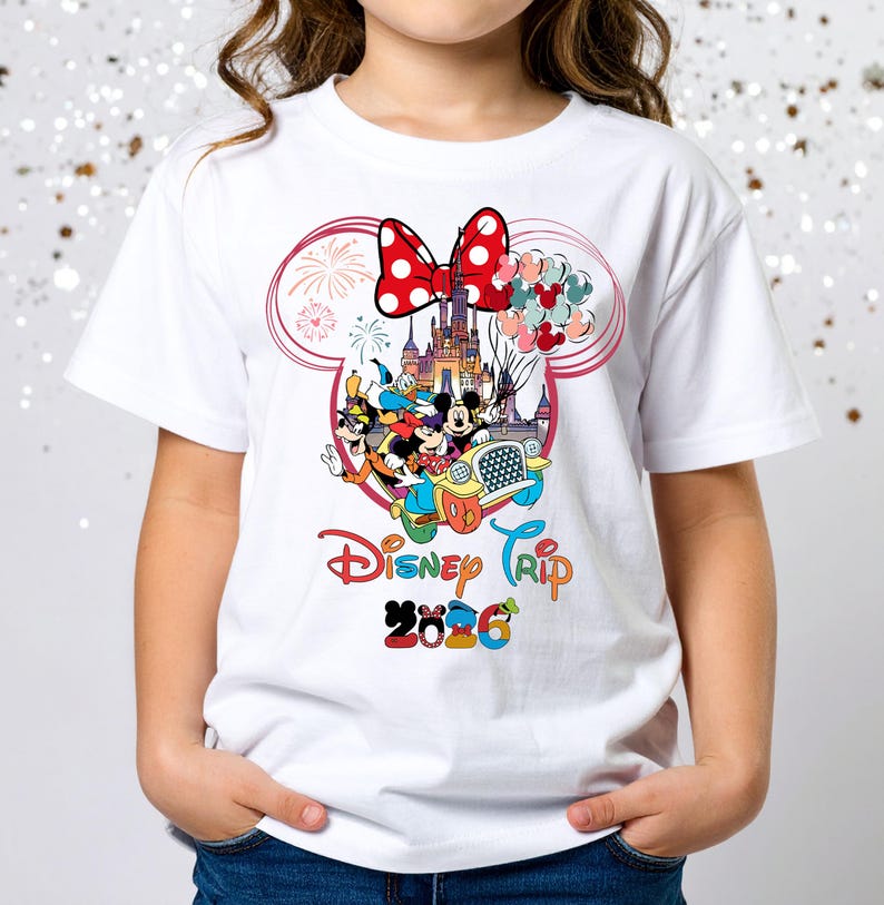 May include: White t-shirt featuring a colorful Disney Trip design. The graphic includes Mickey and Minnie Mouse, Donald Duck, and Goofy in a car, with a castle and fireworks. The design also includes a red bow and the text "Disney Trip 2026".