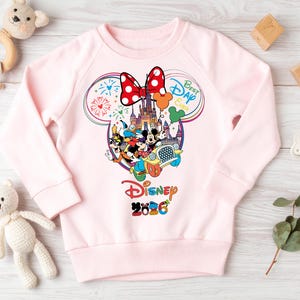 May include: A light pink sweatshirt featuring a colorful Disney graphic. The graphic includes Mickey and Minnie Mouse, a castle, and the text "Best Day Ever" and "Disney 2026". A large red bow with white polka dots is at the top.