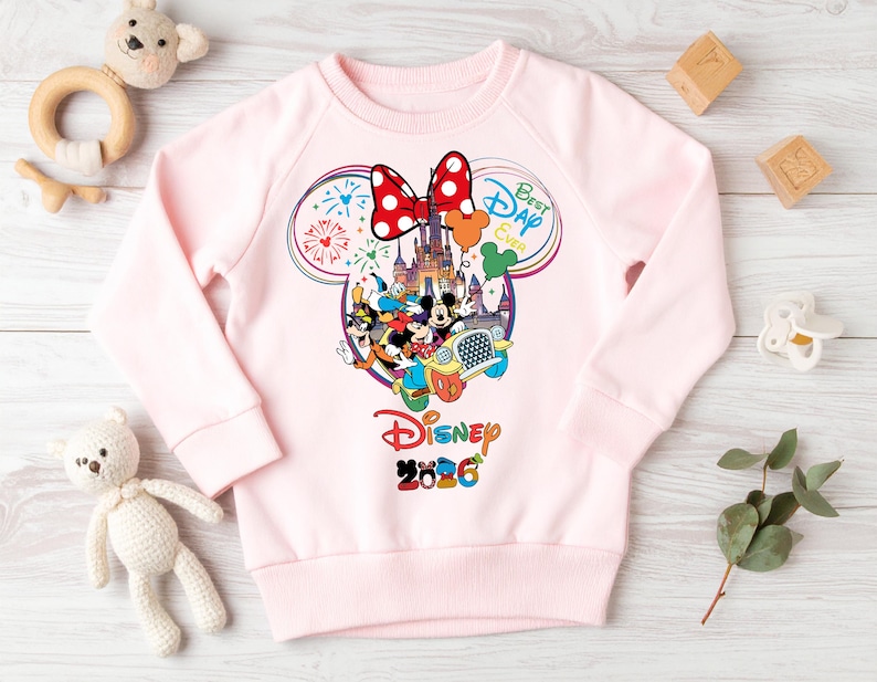 May include: A light pink sweatshirt featuring a colorful Disney graphic. The graphic includes Mickey and Minnie Mouse, a castle, and the text "Best Day Ever" and "Disney 2026". A large red bow with white polka dots is at the top.