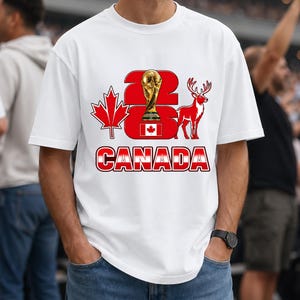 Puede incluir: Camiseta blanca con un dise&ntilde;o gr&aacute;fico que presenta el texto "2026 CANADA" en rojo y blanco, un trofeo dorado, una hoja de arce roja y una silueta de ciervo rojo. La camiseta la lleva una persona.