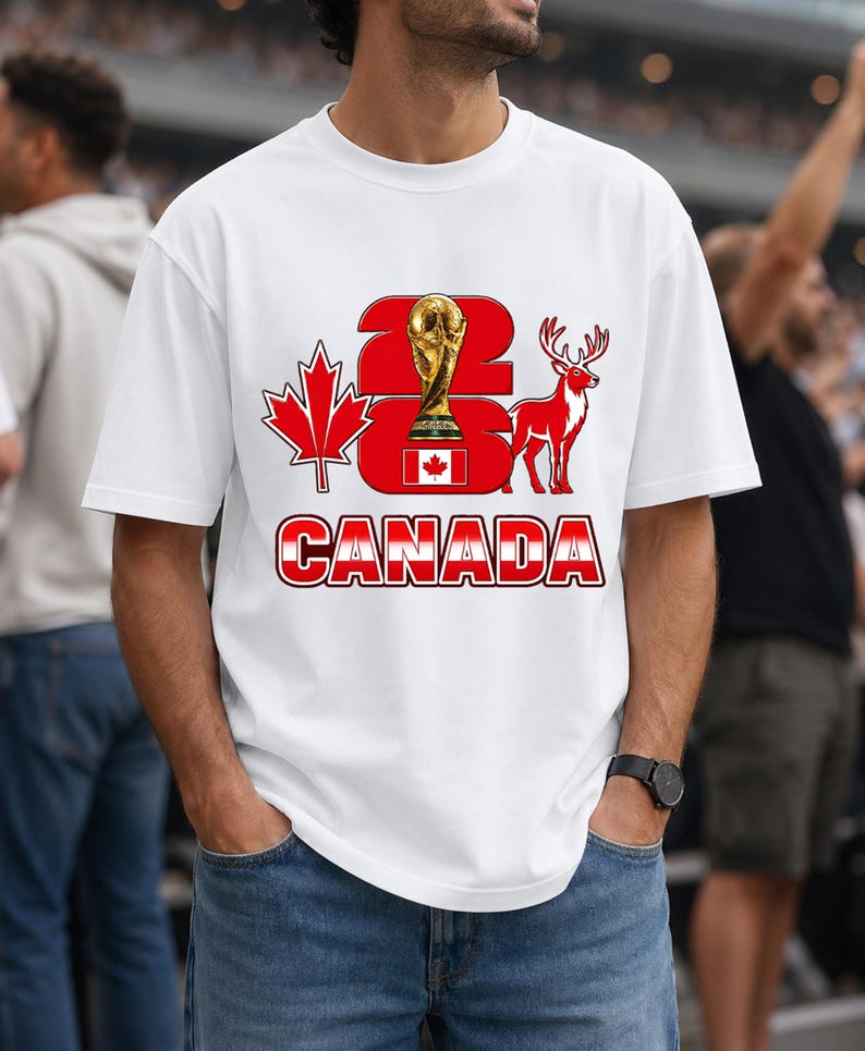 Puede incluir: Camiseta blanca con un dise&ntilde;o gr&aacute;fico que presenta el texto "2026 CANADA" en rojo y blanco, un trofeo dorado, una hoja de arce roja y una silueta de ciervo rojo. La camiseta la lleva una persona.
