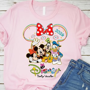 Op de afbeelding: Lichtroze T-shirt met een kleurrijke Disney-afbeelding. Het toont Mickey en Minnie Mouse, Goofy en Donald Duck, met een rode strik met stippen en het jaar 2026. De tekst "Disney Family Vacation" is ook aanwezig.