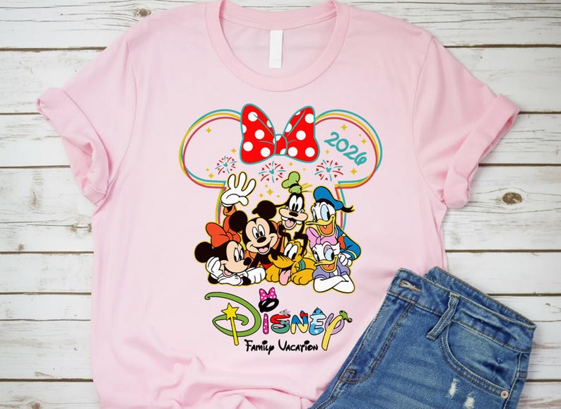 Op de afbeelding: Lichtroze T-shirt met een kleurrijke Disney-afbeelding. Het toont Mickey en Minnie Mouse, Goofy en Donald Duck, met een rode strik met stippen en het jaar 2026. De tekst "Disney Family Vacation" is ook aanwezig.