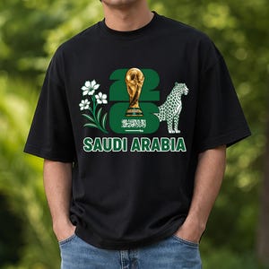 Puede incluir: Camiseta negra con un dise&ntilde;o gr&aacute;fico verde y blanco. El dise&ntilde;o incluye el texto "23", un trofeo dorado, la bandera de Arabia Saud&iacute;, un leopardo blanco y las palabras "SAUDI ARABIA".