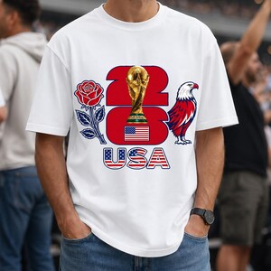 Puede incluir: Camiseta blanca con un dise&ntilde;o que presenta un trofeo dorado, una rosa roja y un &aacute;guila roja, blanca y azul. El dise&ntilde;o incluye el texto "23 USA" en rojo y azul, con la bandera estadounidense.