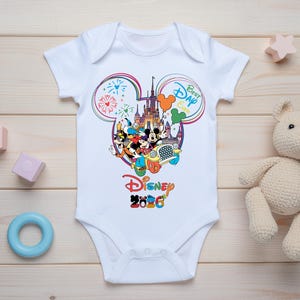May include: White baby onesie with a colorful Disney graphic. The design features Mickey Mouse, Minnie Mouse, and other characters in front of a castle, with the text "Best Day Ever" and "Disney 2020".