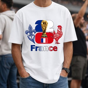 Puede incluir: Camiseta blanca con un dise&ntilde;o gr&aacute;fico que celebra a Francia. El dise&ntilde;o incluye un trofeo dorado, un gallo rojo, la bandera francesa y la palabra "France" en azul. La camiseta la lleva una persona con vaqueros azules.