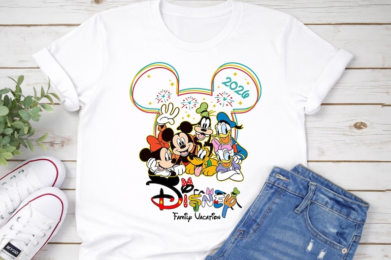 Op de afbeelding: Wit T-shirt met een kleurrijke afbeelding van Disney-personages, waaronder Mickey en Minnie Mouse, Goofy, Donald en Daisy Duck en Pluto. Het ontwerp bevat de tekst "Disney Family Vacation 2026".