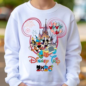 May include: White sweatshirt featuring a colorful Disney graphic. The design includes Mickey Mouse, Donald Duck, and Goofy in a car, with a castle, balloons, and the text "Disney Trip 2026". The graphic is framed by a pink Mickey Mouse ear outline.
