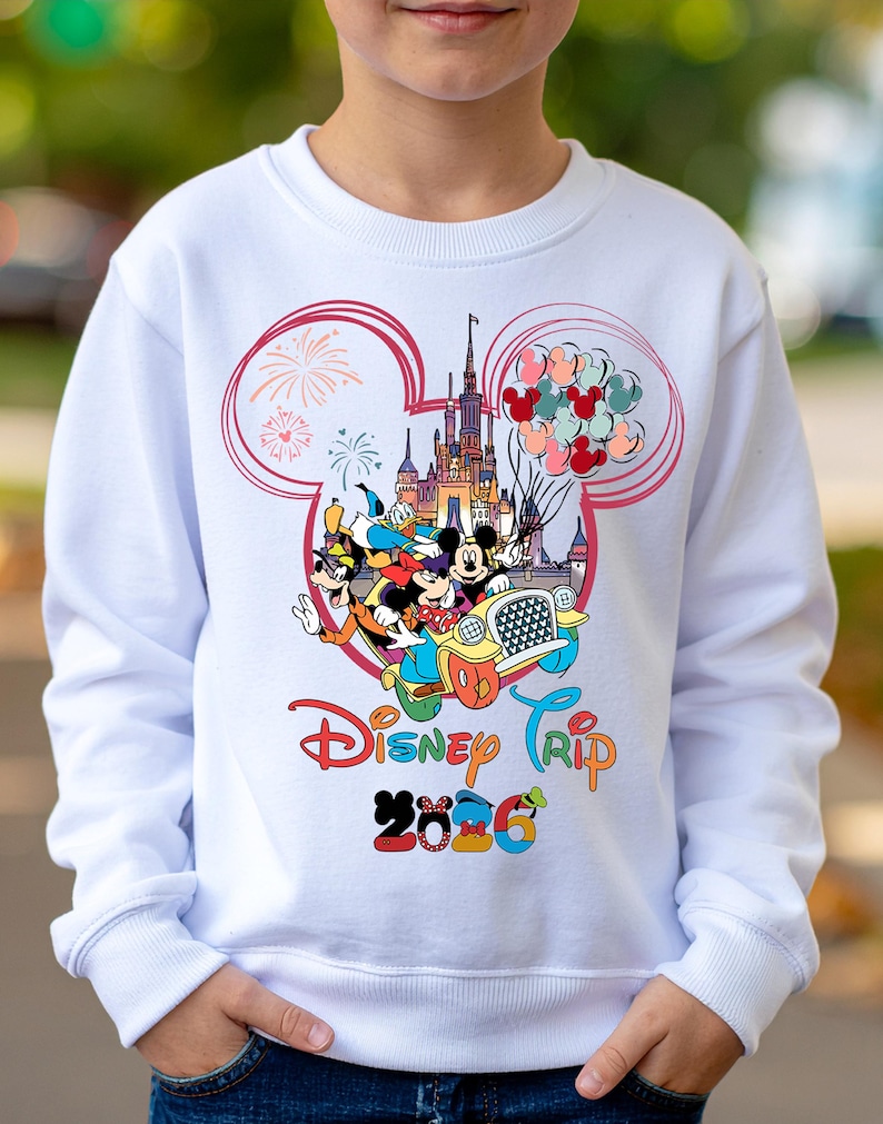 May include: White sweatshirt featuring a colorful Disney graphic. The design includes Mickey Mouse, Donald Duck, and Goofy in a car, with a castle, balloons, and the text "Disney Trip 2026". The graphic is framed by a pink Mickey Mouse ear outline.