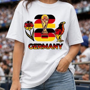 Puede incluir: Camiseta blanca con un dise&ntilde;o gr&aacute;fico que presenta la palabra "GERMANY" en amarillo y rojo, un trofeo, un &aacute;guila y una flor. La camiseta es de manga corta.
