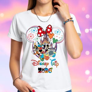 Op de afbeelding: Wit T-shirt met een kleurrijk Disney-ontwerp. Het ontwerp bevat Mickey en Minnie Mouse, een kasteel, vuurwerk en de tekst "Disney Trip 2026". Een rode strik met witte stippen staat bovenaan het ontwerp.