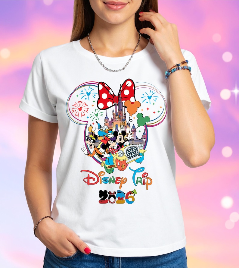Op de afbeelding: Wit T-shirt met een kleurrijk Disney-ontwerp. Het ontwerp bevat Mickey en Minnie Mouse, een kasteel, vuurwerk en de tekst "Disney Trip 2026". Een rode strik met witte stippen staat bovenaan het ontwerp.