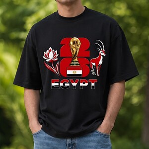 Puede incluir: Camiseta negra con un dise&ntilde;o gr&aacute;fico. El dise&ntilde;o incluye el texto "2026 EGYPT", un trofeo dorado, una flor de loto roja y blanca y una gacela roja y blanca. La bandera egipcia tambi&eacute;n est&aacute; incorporada.