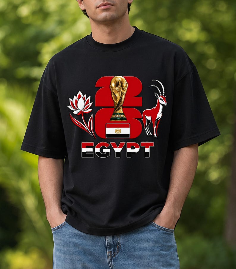 Puede incluir: Camiseta negra con un dise&ntilde;o gr&aacute;fico. El dise&ntilde;o incluye el texto "2026 EGYPT", un trofeo dorado, una flor de loto roja y blanca y una gacela roja y blanca. La bandera egipcia tambi&eacute;n est&aacute; incorporada.