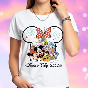 May include: White t-shirt featuring a colorful graphic of Disney characters, including Mickey and Minnie Mouse, Goofy, Donald Duck, and others. The design includes a castle and a red polka-dot bow. The text "Disney Trip 2026" is printed below the characters.