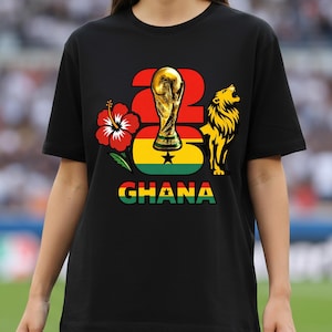 Puede incluir: Camiseta negra con un dise&ntilde;o gr&aacute;fico que incluye el n&uacute;mero 26, un trofeo dorado, una flor de hibisco roja, un le&oacute;n y la palabra "GHANA" en verde, amarillo y rojo. El dise&ntilde;o incorpora los colores de la bandera de Ghana.