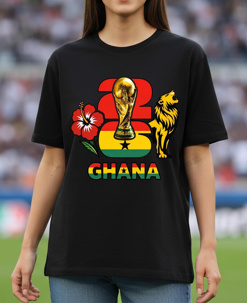 Puede incluir: Camiseta negra con un dise&ntilde;o gr&aacute;fico que incluye el n&uacute;mero 26, un trofeo dorado, una flor de hibisco roja, un le&oacute;n y la palabra "GHANA" en verde, amarillo y rojo. El dise&ntilde;o incorpora los colores de la bandera de Ghana.