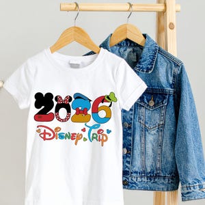 Op de afbeelding: Wit T-shirt met een kleurrijke "2026 Disney Trip" afbeelding. De cijfers zijn ontworpen met Disney-karakterelementen. Een spijkerjasje hangt achter het T-shirt aan een houten rek.